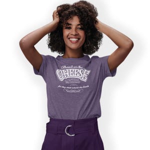 May include: A purple t-shirt with the text "Blessed are the CHEESE MAKERS for they shall inherit the Earth" in white. The shirt is worn by a person with curly dark hair and purple trousers.