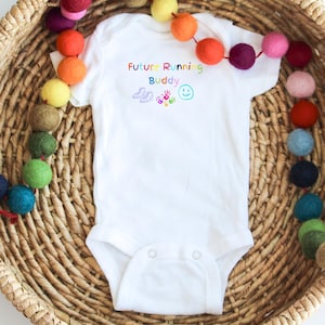 Peut inclure: Combinaison blanche pour bébé avec le texte "Future Running Buddy" en couleurs arc-en-ciel. La combinaison a un dessin d'une paire de chaussures de course, deux empreintes de mains et un visage souriant.