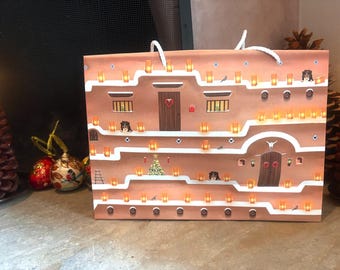 Bolsa de regalo navideña con paredes de adobe cubiertas de nieve y farolitos
