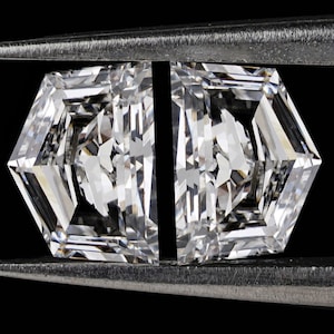 May include: Two clear, octagon-shaped diamonds held in a metal tweezer against a black background.