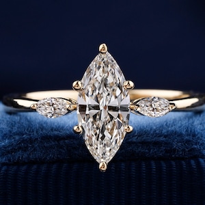 Pu&ograve; includere: Un anello di fidanzamento in oro con un grande diamante a forma di marquise e due diamanti pi&ugrave; piccoli a forma di pera sulla fascia. L'anello &egrave; posto su una scatola di velluto blu.