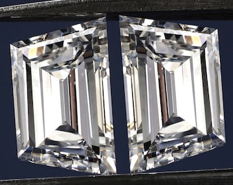 May include: Two clear, square-shaped, emerald-cut diamonds held by a pair of tweezers.