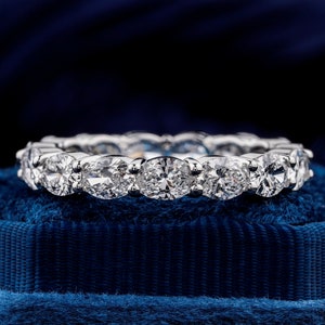 Pode incluir: Uma aliança de eternidade de diamantes com uma fila de diamantes redondos brilhantes engastados em uma banda de ouro branco. O anel é apresentado em um almofada de veludo azul.