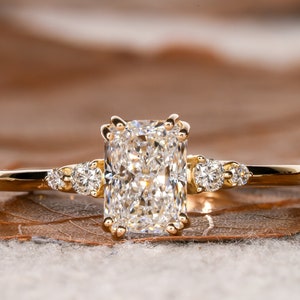 Könnte beinhalten: Ein goldener Verlobungsring mit einem großen, rechteckigen, klaren Diamanten als Mittelstück und drei kleineren, runden, klaren Diamanten auf der Ringschiene.