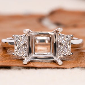 May include: A silver ring setting with three square-shaped prongs for a center stone and two smaller square-shaped prongs on either side for accent stones.