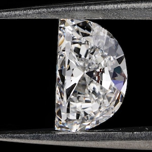 May include: A clear, faceted diamond gemstone held in a pair of silver tweezers against a black background.