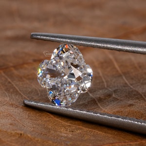 Op de afbeelding: Een heldere, gefacetteerde diamant in de vorm van een klavertjevier, vastgehouden door zilveren pincetten tegen een bruine houten achtergrond.