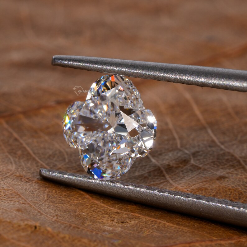 Op de afbeelding: Een heldere, gefacetteerde diamant in de vorm van een klavertjevier, vastgehouden door zilveren pincetten tegen een bruine houten achtergrond.