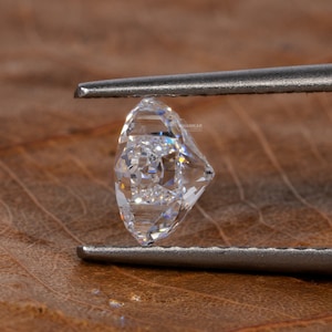 Op de afbeelding: Een heldere, gefacetteerde diamant edelsteen vastgehouden door een pincet tegen een bruine, getextureerde achtergrond.