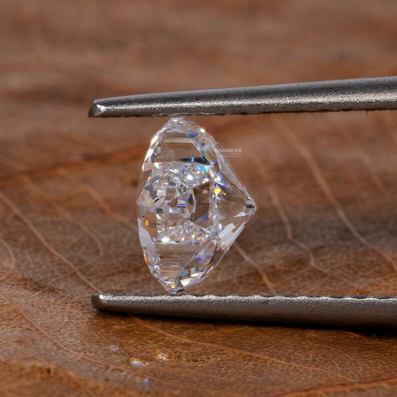 Op de afbeelding: Een heldere, gefacetteerde diamant edelsteen vastgehouden door een pincet tegen een bruine, getextureerde achtergrond.