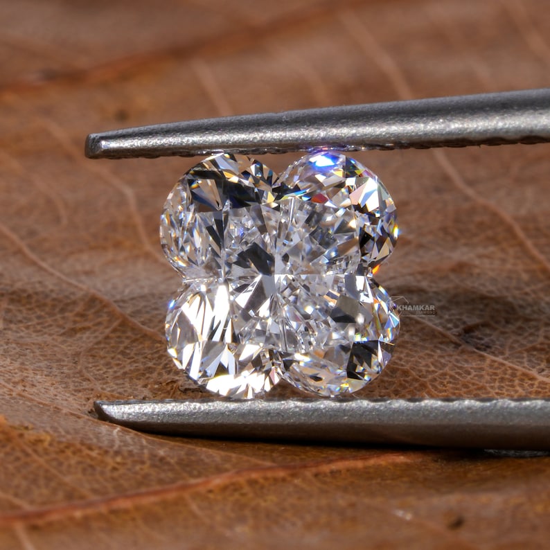 Op de afbeelding: Een heldere, gefacetteerde diamant in de vorm van een klavertjevier, vastgehouden door een zilverkleurige pincet, op een bruin, getextureerd oppervlak.