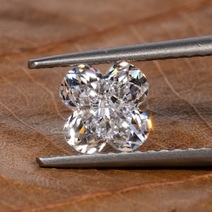 Op de afbeelding: Een heldere, gefacetteerde diamant in de vorm van een klavertjevier, vastgehouden door een pincet tegen een bruine achtergrond.