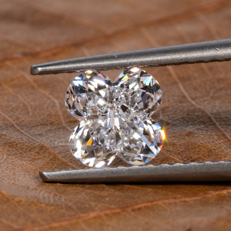 Op de afbeelding: Een heldere, gefacetteerde diamant in de vorm van een klavertjevier, vastgehouden door een pincet tegen een bruine achtergrond.