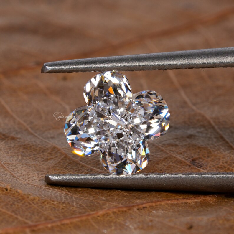 Op de afbeelding: Een heldere, gefacetteerde diamant in de vorm van een klavertjevier, vastgehouden door een pincet tegen een bruine achtergrond.