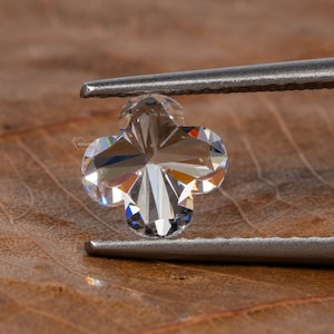 Op de afbeelding: Een heldere, gefacetteerde diamant in de vorm van een klavertjevier, vastgehouden door een pincet tegen een bruine houten achtergrond.