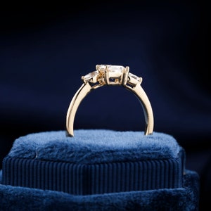 Pu&ograve; includere: Un anello di fidanzamento in oro con tre diamanti rotondi incastonati in un design a tre pietre. L'anello &egrave; su una scatola di velluto blu.