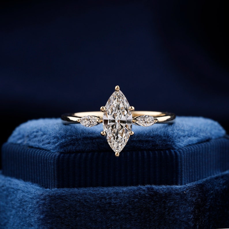 Pu&ograve; includere: Un anello di fidanzamento in oro con un grande diamante a forma di marquise e due diamanti pi&ugrave; piccoli a forma di pera sulla fascia. L'anello &egrave; posto su una scatola di velluto blu.