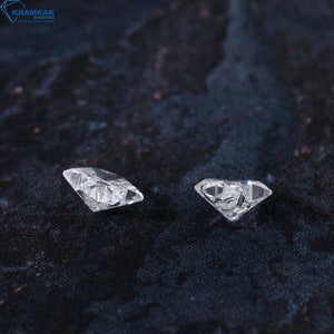 May include: Two clear, faceted diamonds on a dark gray textured surface. The diamonds are different sizes and shapes.
