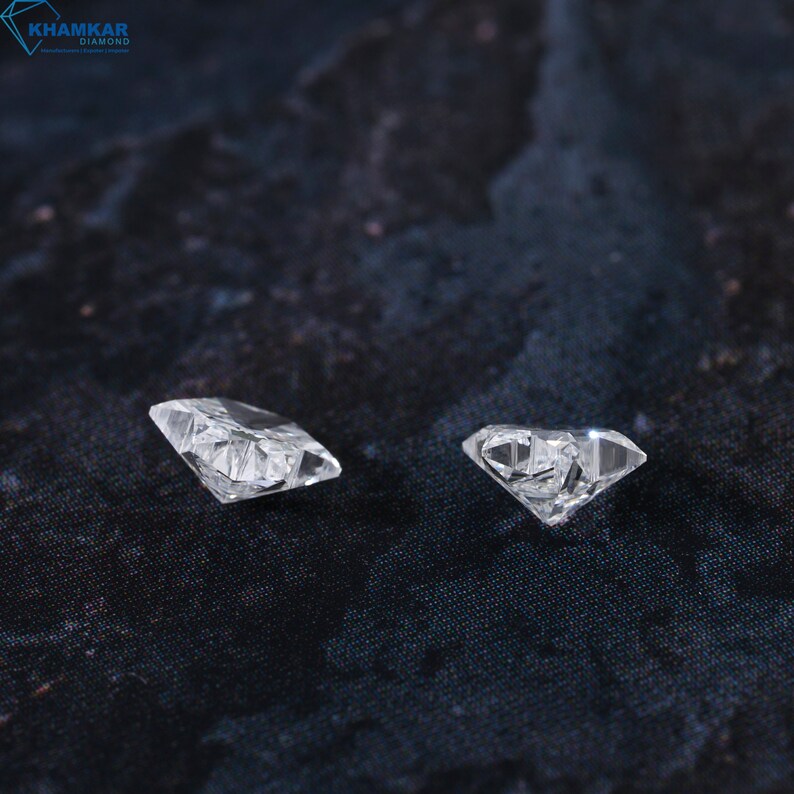 May include: Two clear, faceted diamonds on a dark gray textured surface. The diamonds are different sizes and shapes.
