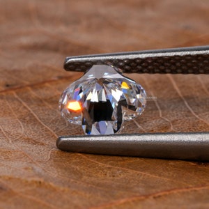 Op de afbeelding: Een heldere, gefacetteerde diamant vastgehouden door een zilveren pincet. De diamant heeft de vorm van een bloem met vijf bloemblaadjes.