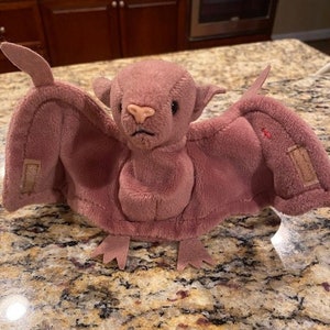 Può includere: Un pipistrello di peluche viola con un naso cucito e grandi orecchie floppy. Il pipistrello è sdraiato su un piano di lavoro marrone e beige.