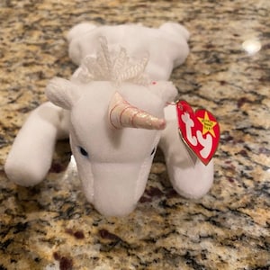 May include: White plush unicorn beanie baby with a pink and white horn, a white sparkly crown, and a red Ty tag with a white star.