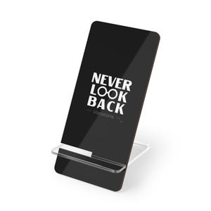 Puede incluir: Un soporte negro para smartphone con el texto "NEVER LOOK BACK - Vacations" en blanco. El soporte tiene una base de acrílico transparente y un borde marrón oscuro.