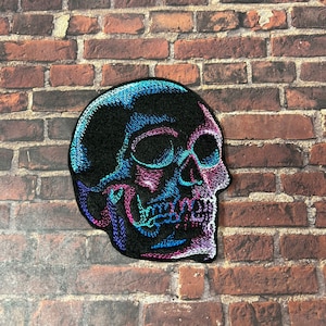 May include: A black embroidered skull patch with blue and pink accents. The skull is facing to the right and has a detailed design.
