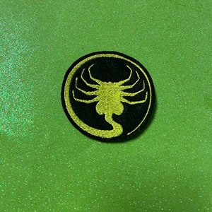 May include: A black and yellow embroidered patch featuring the iconic alien creature from the movie Alien. The patch is circular and has a black background with a yellow alien creature in the center.
