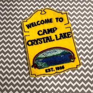 May include: Yellow felt patch with black embroidered text that reads "Welcome to Camp Crystal Lake Est. 1936". The patch features a blue lake with a rainbow above it.