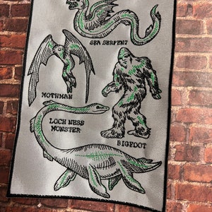 May include: A white patch with black outlines of mythical creatures, including the Loch Ness Monster, Bigfoot, Mothman, and a Sea Serpent. The text "Cryptids of the World" is at the top of the patch.