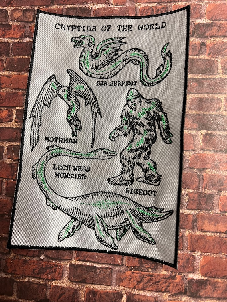 Puede incluir: Un parche blanco con contornos negros de criaturas m&iacute;ticas, incluyendo el monstruo del Lago Ness, Bigfoot, Mothman y una serpiente marina. El texto "Cryptids of the World" est&aacute; en la parte superior del parche.