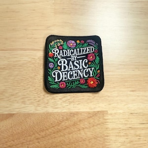 May include: A square black embroidered patch featuring the white text RADICALIZED BY BASIC DECENCY in the center, surrounded by a colorful border of embroidered flowers and green leaves in shades of red, pink, purple, yellow, and orange.