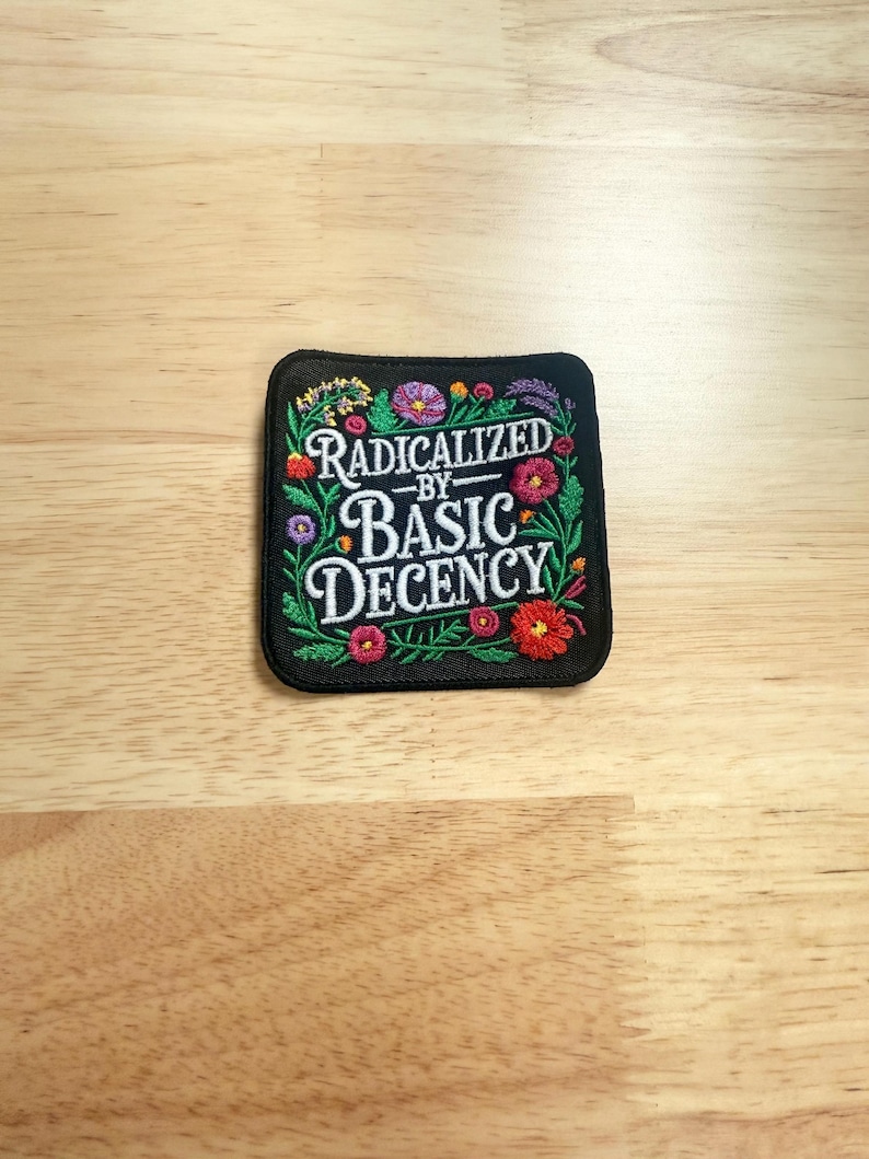 May include: A square black embroidered patch featuring the white text RADICALIZED BY BASIC DECENCY in the center, surrounded by a colorful border of embroidered flowers and green leaves in shades of red, pink, purple, yellow, and orange.