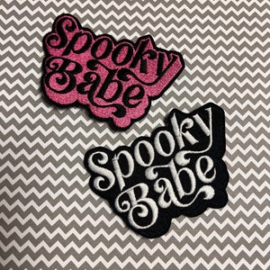 May include: Two embroidered iron-on patches with the text "Spooky Babe" in a retro font. The top patch is pink with black stitching, and the bottom patch is black with white stitching.