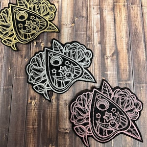 May include: Three embroidered Ouija board patches in gold, silver, and pink. Each patch features a black planchette design with stars, moons, and floral accents. The patches are set against a wooden background.