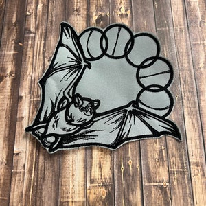 May include: A gray bat patch with black outline and details. The bat is hanging upside down with its wings spread. The patch has a circular design with black circles around the bat.