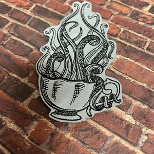 May include: A black and white embroidered patch of an octopus tentacle emerging from a teacup.