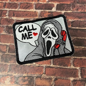 May include: A white and gray embroidered patch featuring a ghost wearing a white mask and holding a red phone. The ghost is saying "Call Me" with a red heart.
