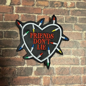 May include: Embroidered patch featuring a black heart shape with the words "FRIENDS DON'T LIE" in red. The heart is outlined with a white string of lights, each with a different colored bulb.