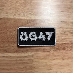 May include: A black rectangular patch with the numbers "8647" embroidered in white. The numbers have a slightly rounded, stylized font and are outlined with a white border. The patch is on a wooden surface.