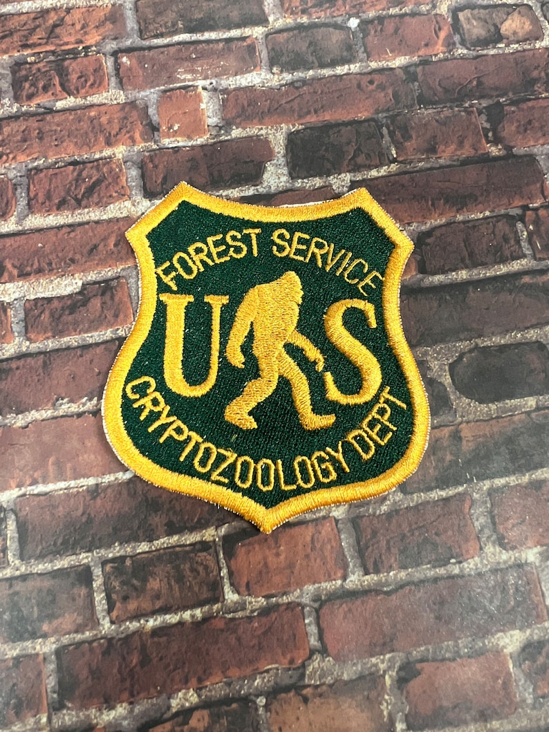 Puede incluir: Un parche bordado en verde y dorado con el texto "Forest Service" y "Cryptozoology Dept." que rodea una silueta estilizada de Bigfoot. El texto "U.S." est&aacute; dentro de la silueta.