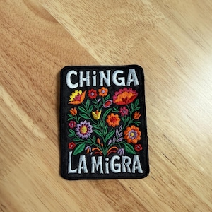 May include: Embroidered patch with the words "CHINGA LA MIGRA" in white, above and below a colorful floral design. The flowers include red, orange, yellow, and purple blooms with green leaves, all on a black background.