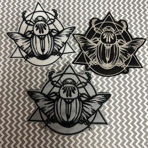 May include: Three embroidered iron-on patches featuring a scarab beetle inside a triangle. The patches are black and white, with the scarab beetle in the center of each patch.