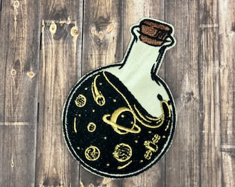 May include: A white glass bottle with a cork stopper filled with a black liquid containing gold planets and stars. The bottle is on a wooden background.