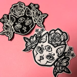 May include: Two black and white embroidered iron-on patches featuring a cat with a third eye and floral designs.