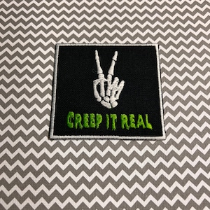 May include: Black iron-on patch with a white skeleton hand making a peace sign and the text "Creep It Real" in green.