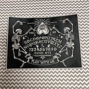 May include: Black and white embroidered patch with a Ouija board design. The board features a heart with an eye, skeletons, and the words "YES", "NO", "GOOD BYE", and "PLAY WITH ME".