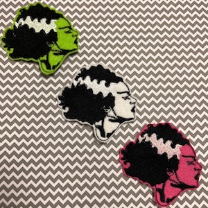 May include: Three embroidered iron-on patches featuring a black and white illustration of a woman with green, white, and pink hair. The woman has a stitched-on scar across her forehead.