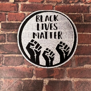 May include: Black Lives Matter patch with three raised fists in black on a white background with a black border.